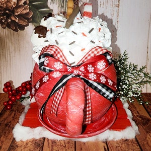 May include: A red ceramic mug with a white rim and handle, decorated with a red and white gingham ribbon and a bow. The mug is filled with white whipped cream, chocolate sprinkles, marshmallows, and candy canes. The mug is sitting on a red wooden coaster.