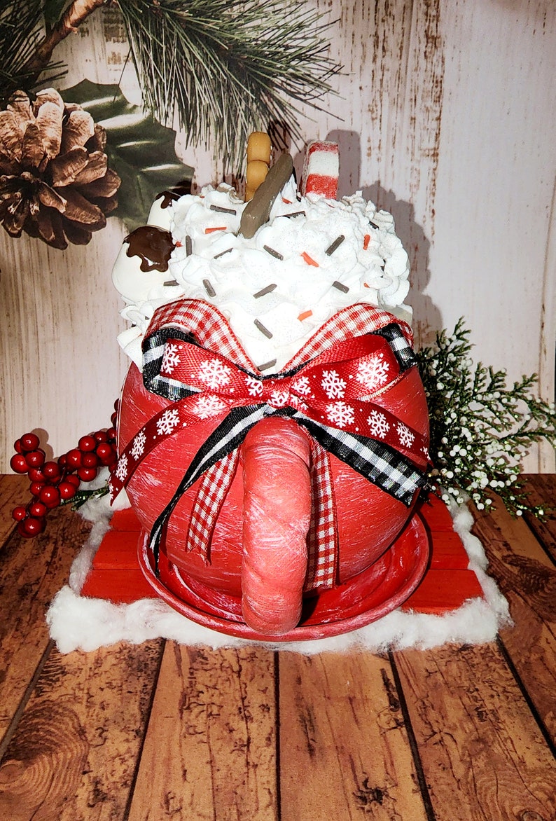 May include: A red ceramic mug with a white rim and handle, decorated with a red and white gingham ribbon and a bow. The mug is filled with white whipped cream, chocolate sprinkles, marshmallows, and candy canes. The mug is sitting on a red wooden coaster.