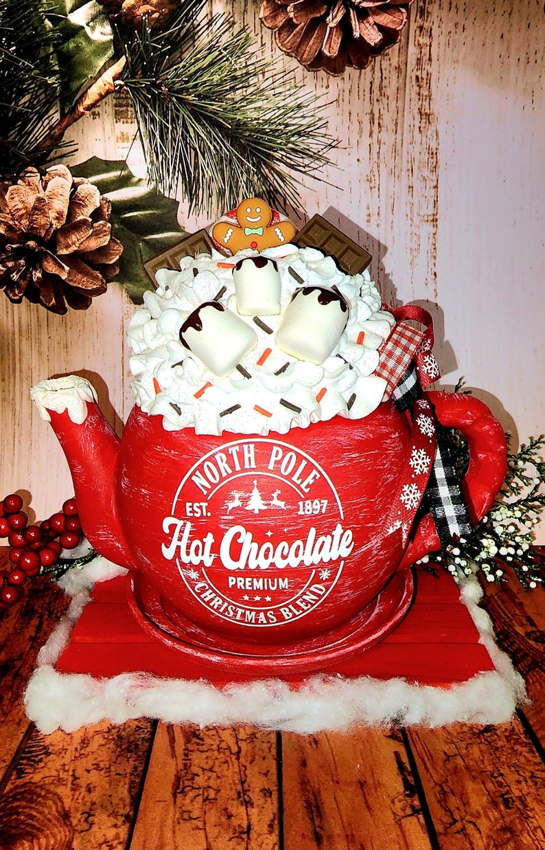May include: Red ceramic teapot with a white and black checkered ribbon, filled with white marshmallows, chocolate sauce, and sprinkles. The teapot has a white label that reads "North Pole Est. 1897 Hot Chocolate Premium Christmas Blend".