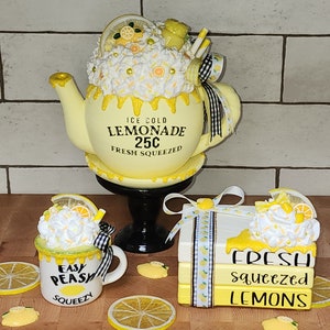 May include: A set of three yellow and white ceramic lemonade themed decor items. A pitcher with a white and yellow frosting design reads "Ice Cold Lemonade 25c Fresh Squeezed". A mug with a white and yellow frosting design reads "Easy Peasy Squeezy". A stack of three books with a white and yellow frosting design reads "Fresh Squeezed Lemons".
