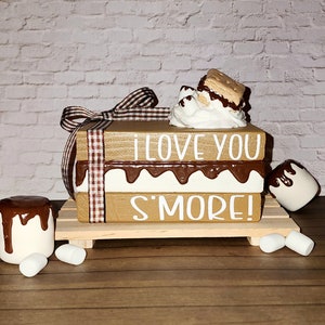 May include: Wooden block sign with the words "I love you s'more!" painted in white. The sign is decorated with a faux chocolate and marshmallow s'more on top.