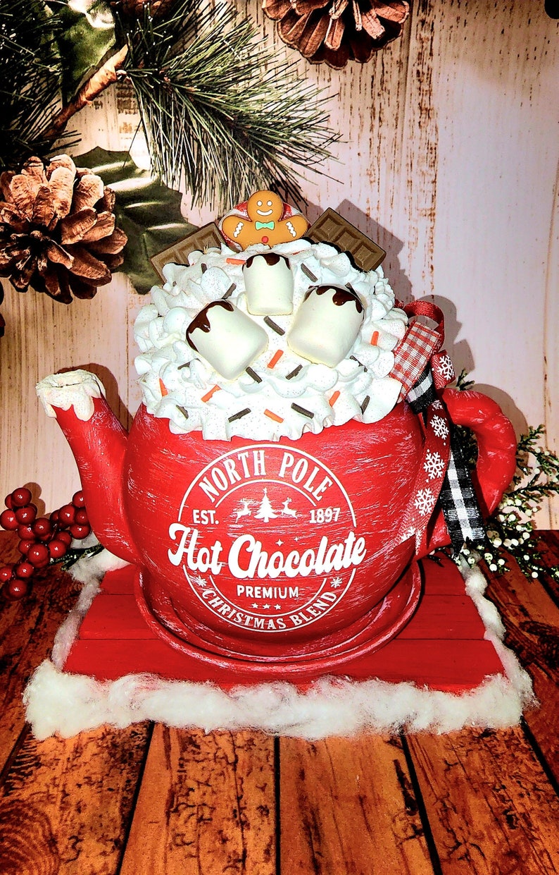 May include: Red ceramic teapot shaped like a mug with white frosting, marshmallows, chocolate sauce, and sprinkles. The teapot has a white label that says "North Pole Est. 1897 Hot Chocolate Premium Christmas Blend". The teapot is sitting on a red wooden base with white faux snow.
