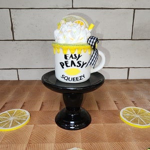 May include: A white ceramic mug with a yellow and white lemon design. The mug has black text that reads "Easy Peasy Squeezy". The mug is sitting on a black pedestal with a lemon slice on the surface.