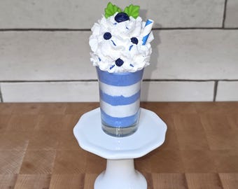 May include: A decorative milkshake with blue and white striped layers in a clear glass. The milkshake is topped with whipped cream, blueberries, and a blue and white striped straw. The glass sits on a white pedestal.