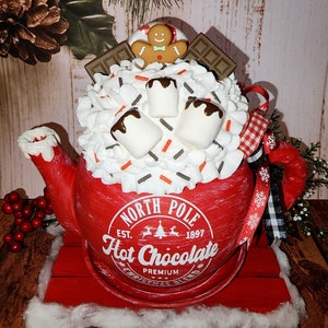 May include: Red ceramic teapot with a white rim and a white label that reads "North Pole Est. 1897 Hot Chocolate Premium Christmas Blend". The teapot is filled with white marshmallows, chocolate sauce, and sprinkles. A gingerbread man cookie sits on top of the teapot.