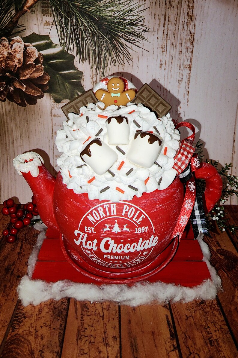 May include: Red ceramic teapot with a white rim and a white label that reads "North Pole Est. 1897 Hot Chocolate Premium Christmas Blend". The teapot is filled with white marshmallows, chocolate sauce, and sprinkles. A gingerbread man cookie sits on top of the teapot.