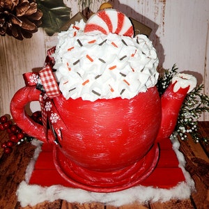 May include: Red teapot with white frosting and red and white candy cane on top. The teapot is sitting on a red wooden base with a white cotton base.
