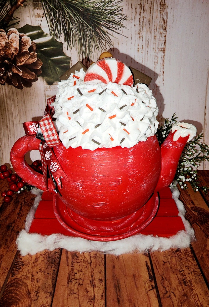 May include: Red teapot with white frosting and red and white candy cane on top. The teapot is sitting on a red wooden base with a white cotton base.