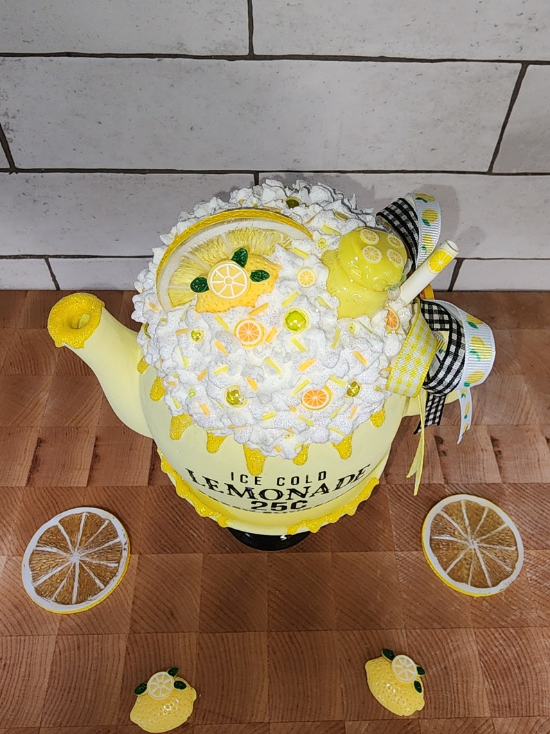 May include: A yellow teapot decorated with white frosting, lemon slices, and sprinkles. The teapot has a black and white checkered ribbon and a straw sticking out of the top. The teapot is labeled "ICE COLD LEMONADE 25&cent;".