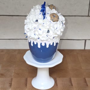 May include: A blue ceramic cup with white frosting, blue sprinkles, and cookie decorations, presented on a white pedestal. The cup has a white drip design. The dessert is on a white, round, pedestal stand.