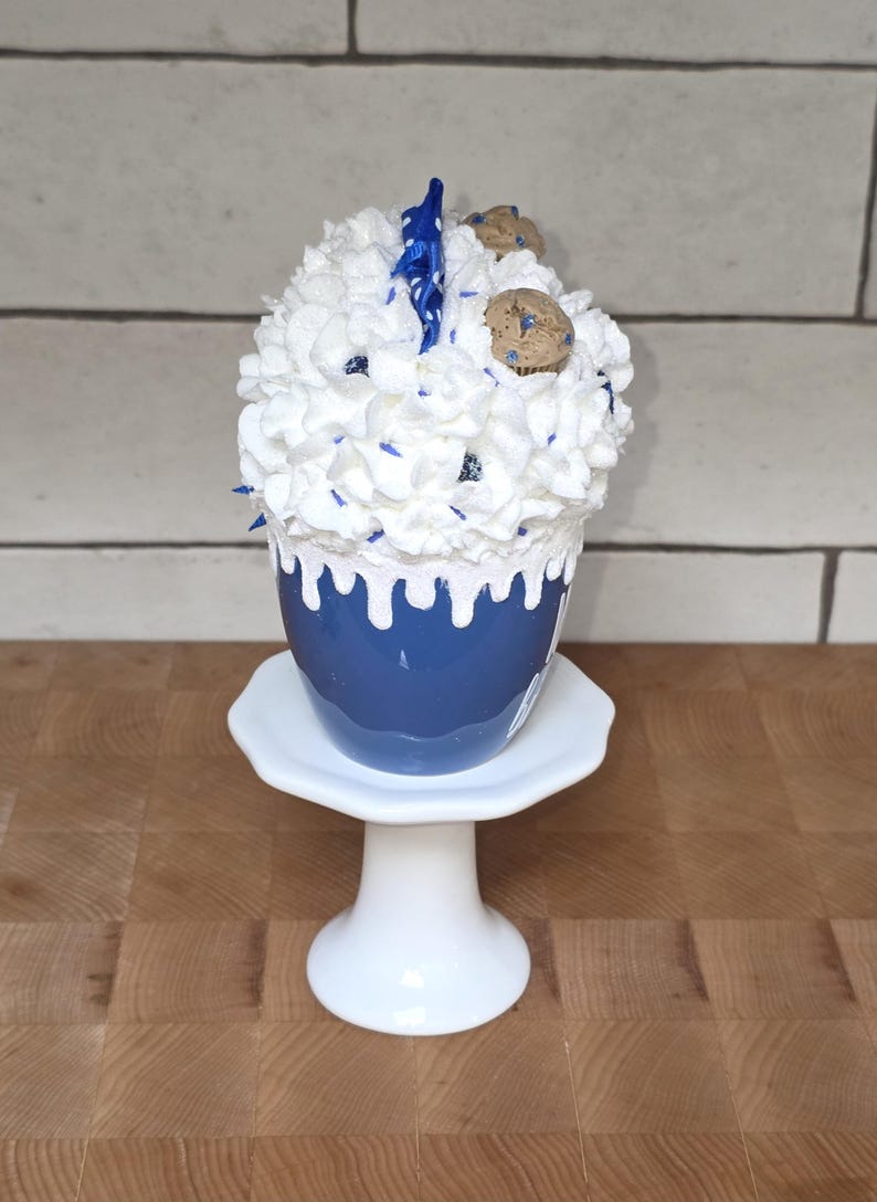 May include: A blue ceramic cup with white frosting, blue sprinkles, and cookie decorations, presented on a white pedestal. The cup has a white drip design. The dessert is on a white, round, pedestal stand.