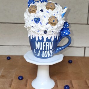 May include: A whimsical blue mug with the text "MUFFIN but LOVE" on a white pedestal. The mug is topped with whipped cream, mini muffins, and blue accents, including a bow and sprinkles. Decorative blueberries are scattered around.