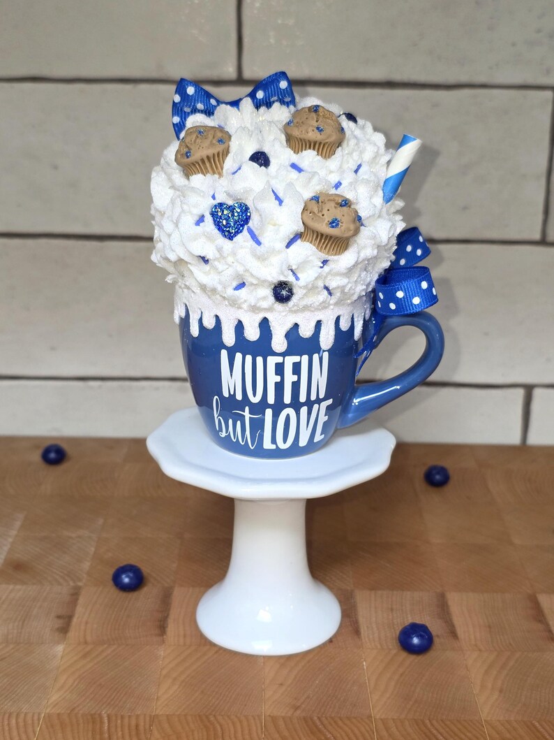 May include: A whimsical blue mug with the text "MUFFIN but LOVE" on a white pedestal. The mug is topped with whipped cream, mini muffins, and blue accents, including a bow and sprinkles. Decorative blueberries are scattered around.