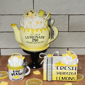May include: A set of three yellow and white ceramic lemonade themed decorations. The first is a teapot with a white and yellow frosting-like topping, a black and white ribbon, and the words "ICE COLD LEMONADE 25C FRESH SQUEEZED". The second is a mug with a white and yellow frosting-like topping, a black and white ribbon, and the words "EASY PEASY SQUEEZY". The third is a stack of three books with a white and yellow frosting-like topping, a black and white ribbon, and the words "FRESH squeezed LEMONS".
