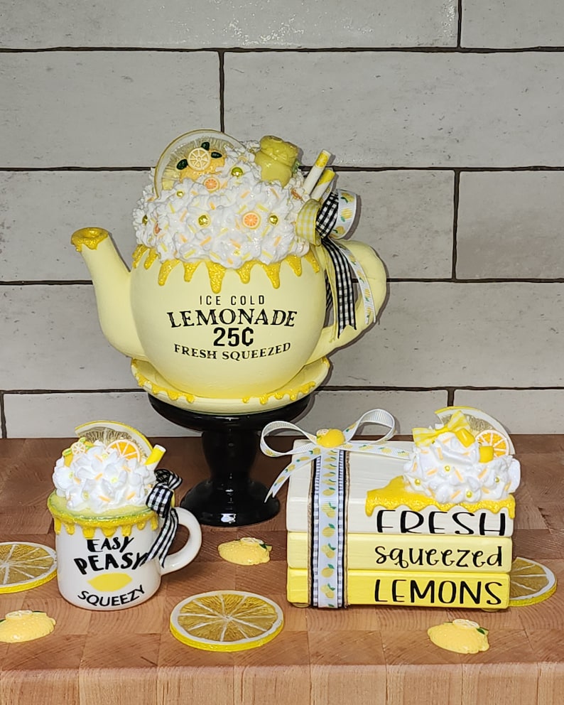 May include: A set of three yellow and white ceramic lemonade themed decorations. The first is a teapot with a white and yellow frosting-like topping, a black and white ribbon, and the words "ICE COLD LEMONADE 25C FRESH SQUEEZED". The second is a mug with a white and yellow frosting-like topping, a black and white ribbon, and the words "EASY PEASY SQUEEZY". The third is a stack of three books with a white and yellow frosting-like topping, a black and white ribbon, and the words "FRESH squeezed LEMONS".