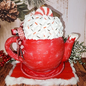 May include: Red ceramic teapot with white frosting and red and white candy on top. The teapot is sitting on a red wooden base with a white fluffy base.