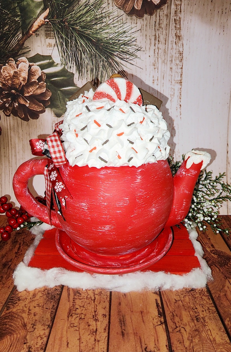 May include: Red ceramic teapot with white frosting and red and white candy on top. The teapot is sitting on a red wooden base with a white fluffy base.