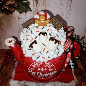 May include: Red ceramic teapot with a white lid and a white, whipped cream-like topping. The teapot is decorated with chocolate syrup, marshmallows, sprinkles, a gingerbread man, and a candy cane. The teapot is sitting on a red wooden sled and has the text "North Pole Est. 1897 Hot Chocolate Premium" on the side.