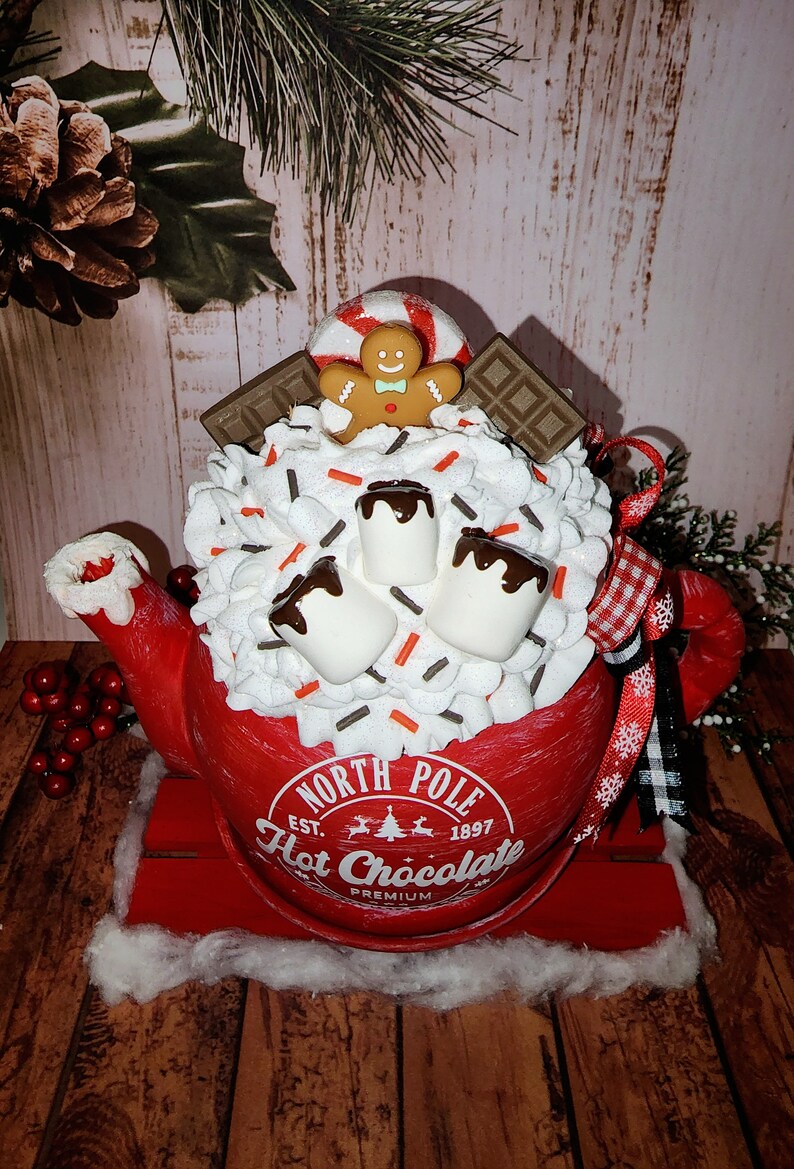 May include: Red ceramic teapot with a white lid and a white, whipped cream-like topping. The teapot is decorated with chocolate syrup, marshmallows, sprinkles, a gingerbread man, and a candy cane. The teapot is sitting on a red wooden sled and has the text "North Pole Est. 1897 Hot Chocolate Premium" on the side.