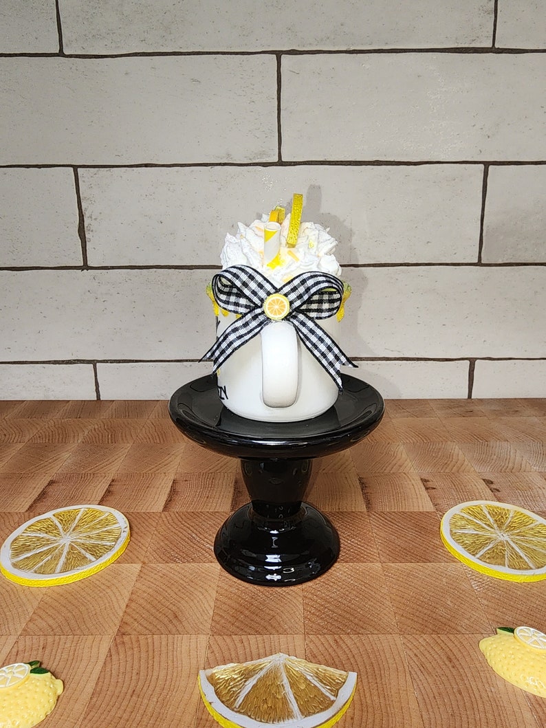 May include: White ceramic mug with a black and white checkered bow, lemon slices, and whipped cream. The mug is on a black pedestal and there are lemon slices on a wooden surface.