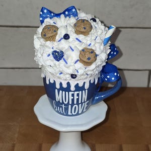 May include: A whimsical blue mug with the text "Muffin but Love" is topped with white whipped cream, miniature muffins, and blue accents. A blue and white polka dot bow and a decorative straw add to the charm. The mug sits on a white pedestal.
