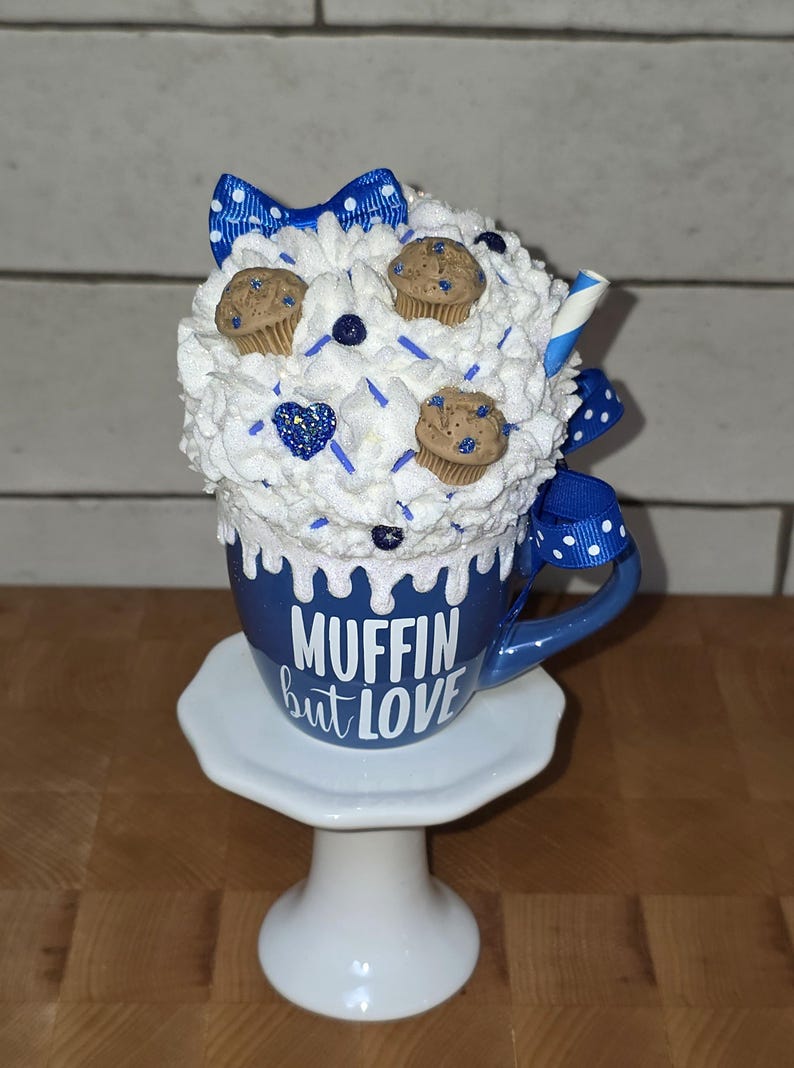 May include: A whimsical blue mug with the text "Muffin but Love" is topped with white whipped cream, miniature muffins, and blue accents. A blue and white polka dot bow and a decorative straw add to the charm. The mug sits on a white pedestal.