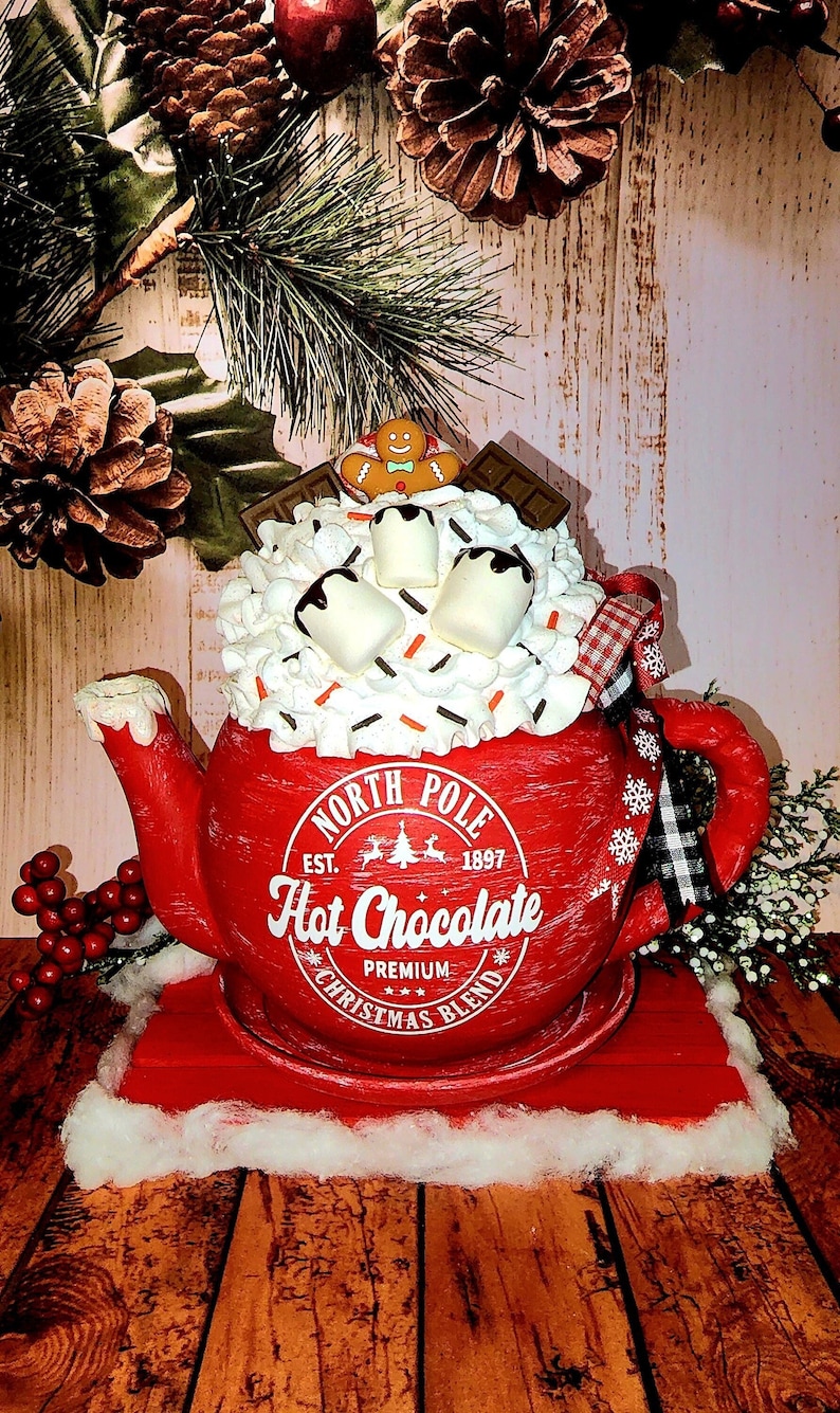 May include: Red ceramic teapot shaped like a mug with white whipped cream, chocolate syrup, marshmallows, and a gingerbread man on top. The teapot has a white label that reads "North Pole Est. 1897 Hot Chocolate Premium Christmas Blend".