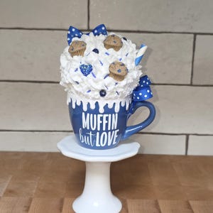 May include: A blue mug with the text "MUFFIN but LOVE" on a white pedestal. The mug is topped with white whipped cream, miniature muffins, blue sprinkles, and a blue and white polka dot bow. A blue and white striped straw is also visible.