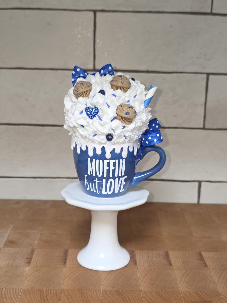 May include: A blue mug with the text "MUFFIN but LOVE" on a white pedestal. The mug is topped with white whipped cream, miniature muffins, blue sprinkles, and a blue and white polka dot bow. A blue and white striped straw is also visible.