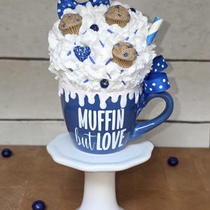 May include: A whimsical blue mug with the text "MUFFIN but LOVE" is topped with white whipped cream, miniature muffins, and a blue bow. The mug sits on a white pedestal, with scattered blueberries.