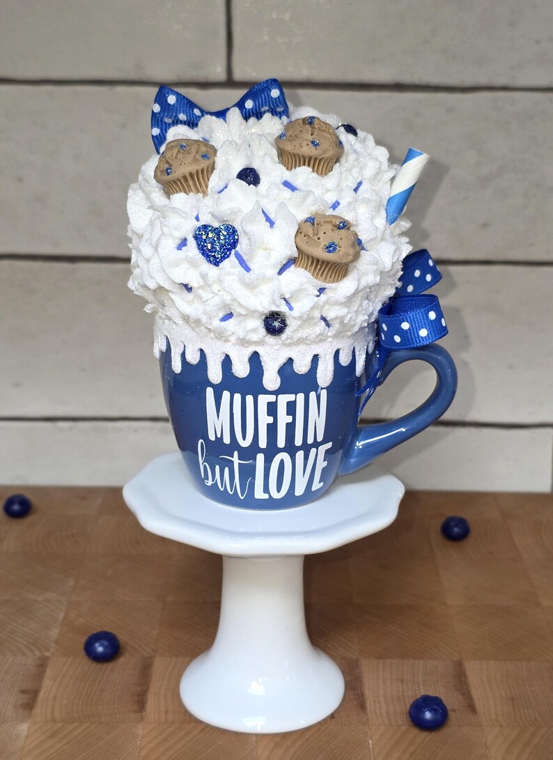 May include: A whimsical blue mug with the text "MUFFIN but LOVE" is topped with white whipped cream, miniature muffins, and a blue bow. The mug sits on a white pedestal, with scattered blueberries.