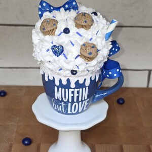 May include: A blue mug with the text "MUFFIN but LOVE" on a white pedestal. The mug is topped with whipped cream, miniature muffins, and a blue bow. A blue and white straw is visible.