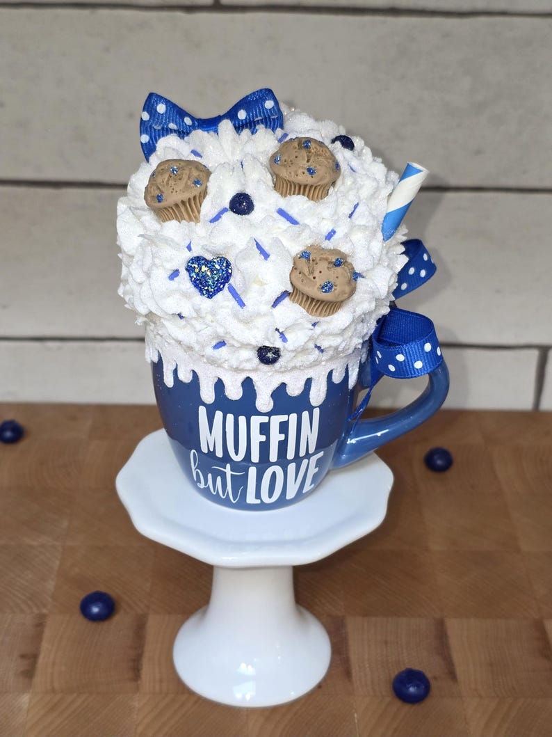 May include: A blue mug with the text "MUFFIN but LOVE" on a white pedestal. The mug is topped with whipped cream, miniature muffins, and a blue bow. A blue and white straw is visible.