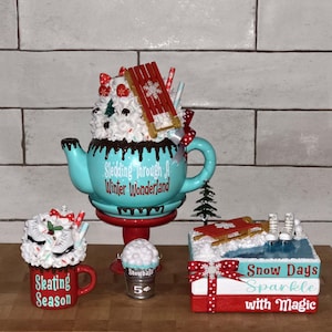Puede incluir: Conjunto decorativo de invierno. Incluye una tetera turquesa con el texto "Sledding Through A Winter Wonderland", una taza roja con "Skating Season", un cubo de bolas de nieve y una pila de libros con "Snow Days Sparkle with Magic".