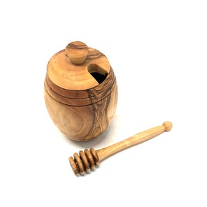 Könnte beinhalten: Ein Honigtopf aus Holz mit Deckel und einem Holzhoniglöffel. Der Honigtopf ist aus Olivenholz gefertigt und hat eine natürliche Oberfläche. Der Honiglöffel ist ebenfalls aus Olivenholz und hat ein Spiraldesign.