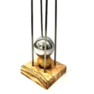 Op de afbeelding: Een zilveren metalen bal die in een metalen frame hangt, rustend op een vierkante houten basis met een natuurlijk houtnerfpatroon.