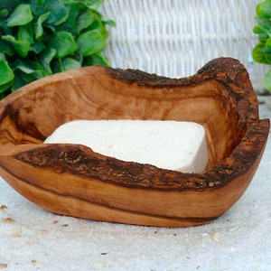 Könnte beinhalten: Handgefertigte Seifenschale aus Olivenholz mit rustikalem Design. Sie enthält ein rechteckiges Stück weiße Seife. Die Holzmaserung und die Rinde sind sichtbar, was dem Ganzen eine natürliche Ästhetik verleiht.