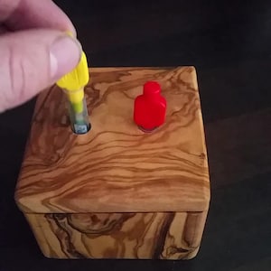 Könnte beinhalten: Eine Holzkiste mit Deckel, mit einem natürlichen Holzmaserungsmuster. Ein gelbes und klares Objekt wird in ein Loch in der Kiste gesteckt. Ein rotes Objekt liegt oben auf der Kiste.