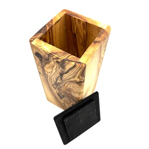May include: A tall, square wooden container with a natural wood grain pattern. The container has a dark, rectangular lid. The wood has a light brown color with darker brown swirls and patterns.