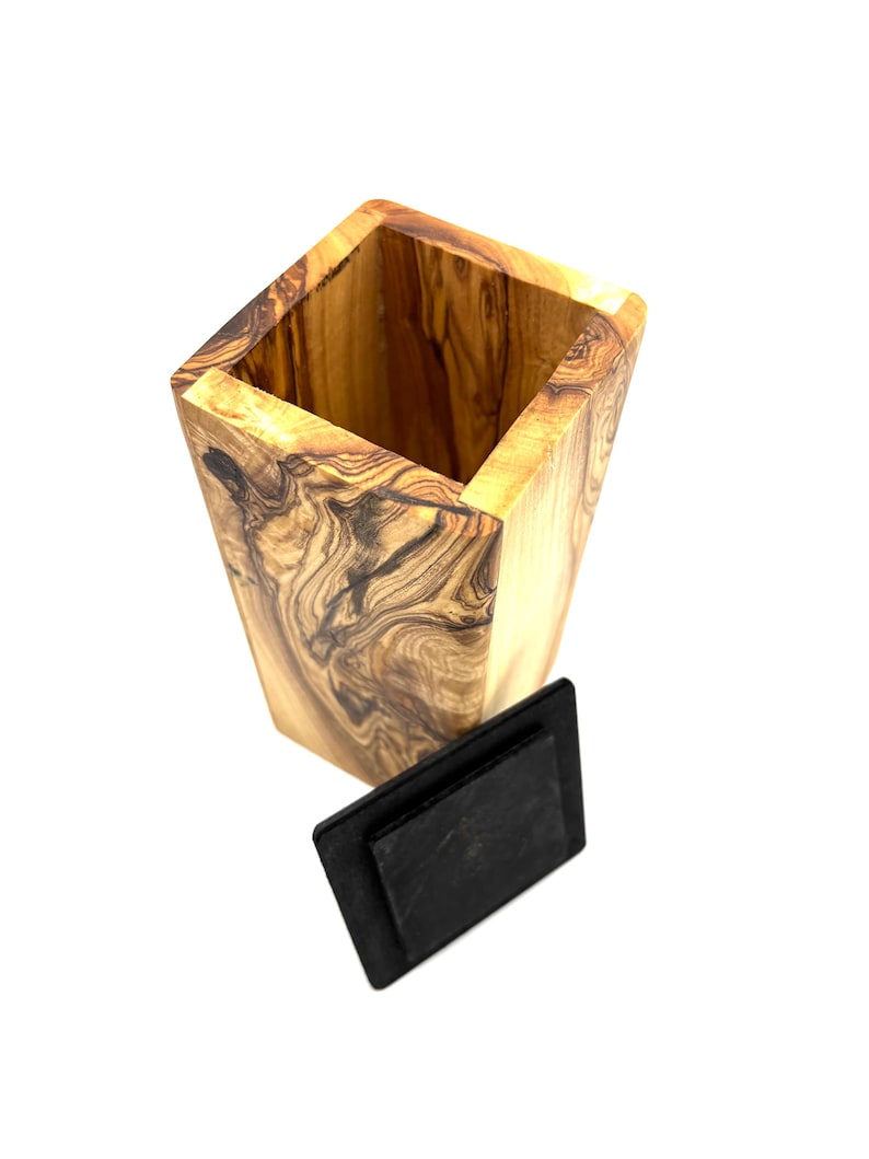 May include: A tall, square wooden container with a natural wood grain pattern. The container has a dark, rectangular lid. The wood has a light brown color with darker brown swirls and patterns.