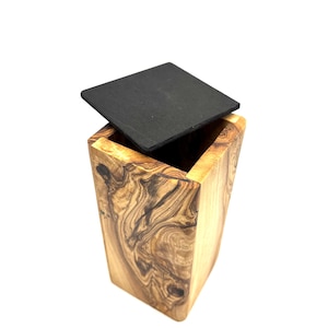 May include: A wooden box with a square, black lid. The box is made of olive wood, showcasing a natural grain pattern in shades of brown and tan. The lid is slightly ajar, revealing the box's interior.