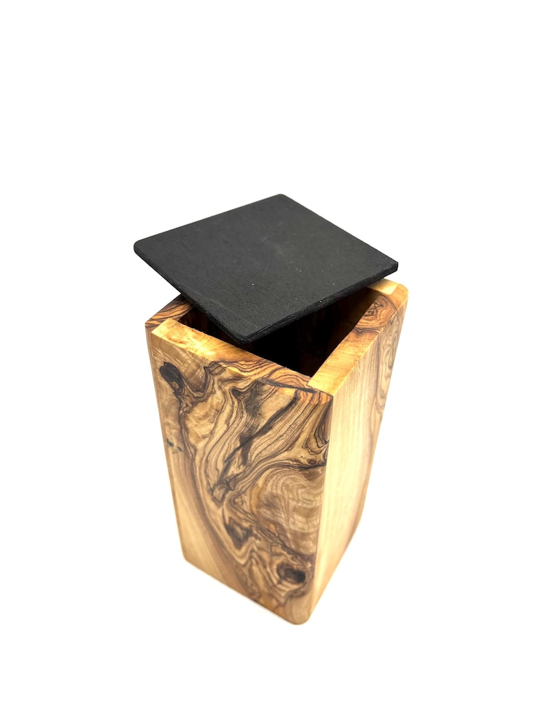 May include: A wooden box with a square, black lid. The box is made of olive wood, showcasing a natural grain pattern in shades of brown and tan. The lid is slightly ajar, revealing the box's interior.