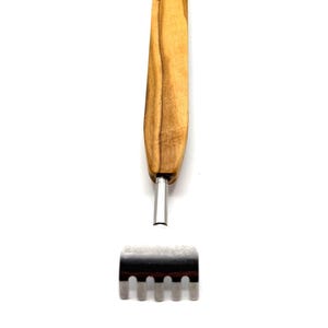 Puede incluir: Un rascador de espalda con mango de madera y cabezal rascador de acero inoxidable. El mango es de color marrón claro y el cabezal tiene cinco púas redondeadas. La herramienta está diseñada para uso personal.