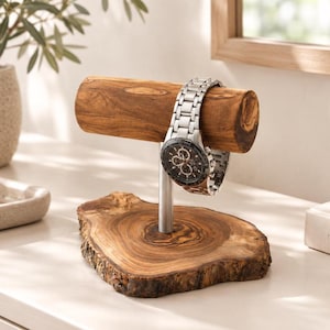 Op de afbeelding: Een horlogestandaard met een horizontale cilindrische houten balk, ondersteund door een verticale zilverkleurige metalen stang op een natuurlijke houten voet met boomschorsrand. Een zilverkleurig analoog horloge met metalen schakelband.