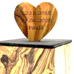 May include: A wooden heart-shaped keepsake with the dates 12.12.2018 and 27.06.2025, and the name Paula, engraved on it. The heart sits atop a dark, rectangular base, which is placed on a wooden box with a unique grain pattern.