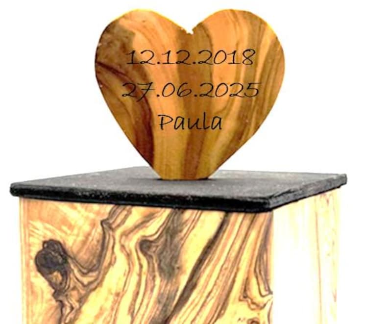 May include: A wooden heart-shaped keepsake with the dates 12.12.2018 and 27.06.2025, and the name Paula, engraved on it. The heart sits atop a dark, rectangular base, which is placed on a wooden box with a unique grain pattern.