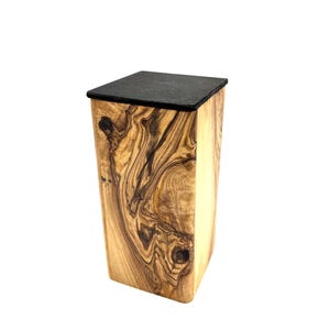 May include: A wooden box with a black top. The box is made of light brown wood with dark brown grain patterns. The box has a square shape and is slightly tapered.