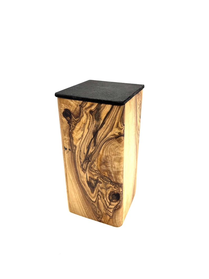 May include: A wooden box with a black top. The box is made of light brown wood with dark brown grain patterns. The box has a square shape and is slightly tapered.
