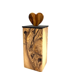 May include: A wooden urn with a heart-shaped lid. The urn is rectangular with a natural wood grain pattern. The lid is black, and the heart is made of the same wood as the urn. The urn is likely for storing ashes.