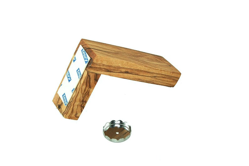 Puede incluir: Un abridor de botellas de madera con un patr&oacute;n de veta de madera marr&oacute;n y negro. El abridor est&aacute; fijado a una pared con cinta adhesiva. Un tap&oacute;n de botella plateado est&aacute; sobre la superficie debajo del abridor.